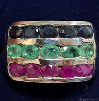 Front face close-up showing three rows of faceted oval black sapphires, green emeralds, and red rubies set in wide silver band.