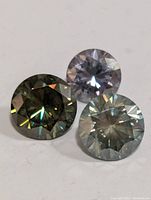 Close-up photo of three round-cut faceted moissanite stones with diamond-like brilliance, different subtle colors including clear and tinted.