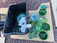 Glassware assortment in blue and green shades arranged beside black plastic tote container, showing variety of bowls, plates, and pitchers.