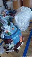 Looney Tunes cookie jar with Taz and Bugs Bunny alongside white ceramic turkey figurine on wood surface with garage items in background.