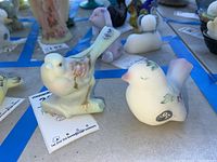Photo showing two bird figurines placed on a table with blue tape lines, each with visible signature labels and floral painted details.