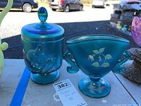 Photo showing both blue glass items side by side on a table outdoors, including the lidded candy dish with bird motif and the vase with floral painting.