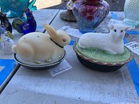 Both bunny and lamb lidded dishes on table with other decorative items in background.