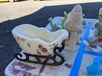 Photo showing white frosted glass sleigh with floral and holly berry decorations and metal runners next to Santa figurine on a table surface outdoors.