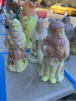Two custard satin glass Santas with pink floral decorations in foreground, additional Santas behind.