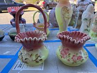 Two Fenton glass pieces, a small basket with handle and a ruffled vase, both painted with floral designs and pink ruffled rims.