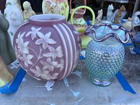 Two vases side by side on table among other items. Left purple floral embossed vase and right iridescent hobnail Fenton vase with ruffled edge.