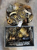Two boxes filled with assorted vintage metal lamp parts including bases, shades, rings, and wiring components laid out on a garage floor.