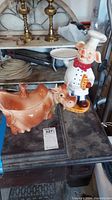 Photo showing orange ceramic cow-shaped cookie jar with small deer figure and pig cookie jar with tray on table with other objects in background.