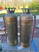 Two vintage metal fire extinguishers side by side outdoors, showing full body and attached hoses