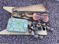 Damaged violin inside its hard case resting on cardboard with vintage metal tin and metal rain chains beside it on a rocky ground.