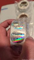 Hand holding a roll of round holographic thank you stickers sealed in plastic. Stickers have texts including 'Happy Mail' and 'order' visible in shiny font.