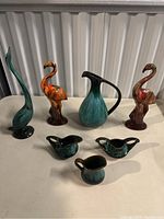 Seven ceramic pieces on a table including bird figurines and pitchers with various glaze colors.