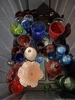Top view of a collection of colorful glassware in various colors including blue, red, green, and clear, showing many drinking glasses, bowls, and jars.