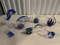 Overall view of nine glass dolphin and fish figurines placed on a white surface, showing different shapes, finishes and colors of blue and clear glass with multicolored accents.