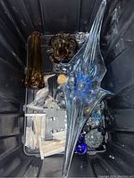 Top-down view of assorted glass and marble pieces in plastic bin including blue elongated glass sculpture, amber bowl, and marble pieces