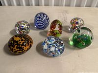 Seven assorted glass paperweights in various colors and designs arranged on a white surface.