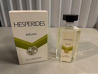 Photo showing the Hesperides Jean Menouns cologne bottle next to its original box. The bottle is clear glass with a black cap and pale yellow liquid inside. The white box displays the brand name and has green and gold accents.