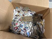 Cardboard box filled with numerous postage stamps on small pieces of paper, various colors and designs visible.