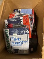 Box containing multiple new men's crew neck t-shirts in original packaging. Various colors and brands visible.