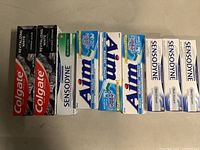 Photo showing nine toothpaste boxes of various brands including Colgate, Sensodyne, Crest, and Aim arranged side by side on a flat surface.