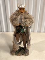 Front view of Viking figurine showing detailed face, silver horned helmet, brown pants, green tunic, and large fur cloak draped over shoulders.