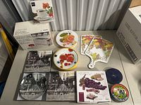 Overview of lot on table including decorative plates, ceramic cheese boxes, boxed serving dishes, and cutting boards