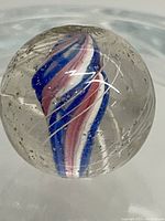 Close-up photo showing the clear glass marble with a red, white, and blue swirl core and visible internal bubbles.