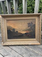 Front view of the antique landscape oil painting with ornate wooden frame outdoors on wooden deck railing.