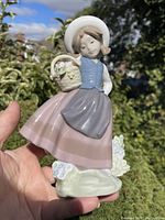 Figurine held outside showcasing full body view with hat, blue vest, pink skirt, gray apron, and basket.