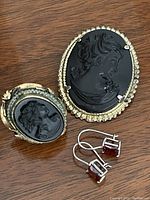 Top-down image showing black cameo ring, black cameo pin, and garnet color earrings laid out.