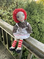 Full body image of the Raggedy Anne doll seated outdoors on a wooden railing with greenery in the background. Doll is wearing original floral dress, white apron, striped stockings, and black shoes.
