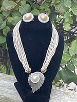 Front view of the necklace on black stand alongside matching earrings, showing multi-strand faux pearls and large pendant with pearl and rhinestones.