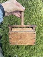 Buttton press held upright by hand showing the flat pressing board with handle and nails.