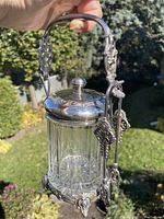 Overall view of pickle cruet held by handle showing full piece with jar, lid, stand and tongs