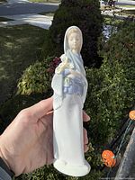 Full view of the 10-inch tall porcelain figurine of a woman in cloak holding flowers, showing overall condition and size relative to hand.