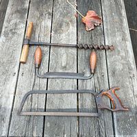 Three antique hand tools laid flat on wooden surface: a T-handle auger drill, a bow saw, and a coping saw.