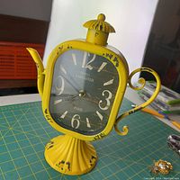 Front angle of yellow teapot shape clock showing distressed paint and clock face with 'Hotel Lancaster Paris'
