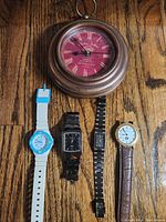 Overview of round wall clock and four wristwatches — white-blue Casio, black KARL, black rectangular, and round gold-tone watch with brown strap