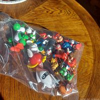 Plastic bag with assorted Super Mario character figurines including Mario, Yoshi, Goomba, Koopa Troopa, and Blooper laid on wooden surface