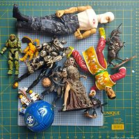 Photo shows a collection of various action figures and toys including GI Joe, Halo Spartan, Transformers-like robots, Bakugan figure, fantasy themed figures, and a blue helmet accessory arranged on a cutting mat.