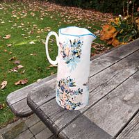 Tall ceramic jug with hand-painted floral design on a wooden outdoor table.