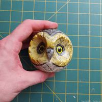 Hand holding small spherical stone owl showing detailed facial features, large yellow eyes, and carved feather pattern.