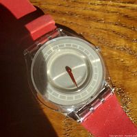 Close-up of the Swatch watch face showing transparent case, silver dial and red hour hand.