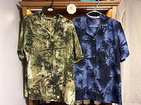 Two men's button down short sleeve dress shirts hanging on hangers, one green and black abstract print and one navy blue with black palm leaf pattern, both size 14.