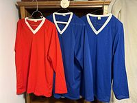 Three long-sleeve jerseys hanging on hangers: one red size L and two blue size XL, all with white V-neck trim.