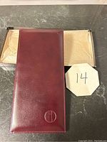 Closed long maroon leather feel card holder showing the smooth front exterior and embossed circle logo.