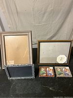 Five picture frames of different sizes and styles placed on a black surface with a beige backdrop, include small decorative tiles