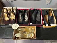 Top-down view of five pairs of women's shoes in their boxes: maroon heels, two pairs of black heels, black loafers, and gold heels.