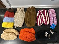 Photo shows eight knitted slippers arranged in two rows on a black countertop: five new pairs on top row and three worn pairs on bottom row. Various colors and styles including striped, solid, and fuzzy.